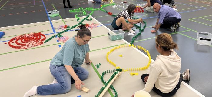 Teilnehmer einer Firmen-Teamchallenge bauen gemeinsam ein buntes Logo aus tausenden Dominosteinen in einer Eventhalle