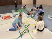 Teilnehmer einer Firmen-Teamchallenge bauen gemeinsam ein buntes Logo aus tausenden Dominosteinen in einer Eventhalle